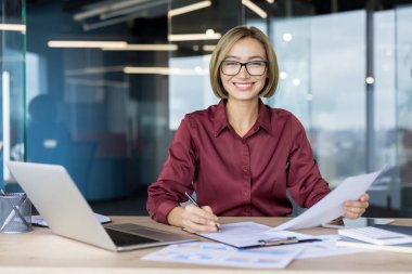 Confident asian business woman smiling and looking at camera while working with papers and laptop at a modern office desk, representing productivity and corporate success