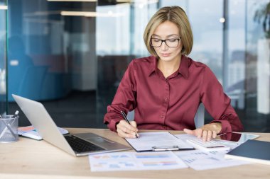 Gözlük takan, mali durumu dengeleyen, belgeleri yöneten ve modern ofis ortamında dizüstü bilgisayarla masrafları hesaplayan profesyonel bir kadın.