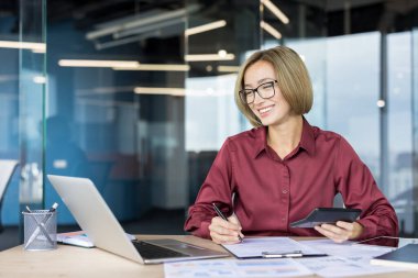 Smiling businesswoman in glasses at a modern office desk, working on laptop and paperwork with a calculator, focused on budgeting, planning and professional success