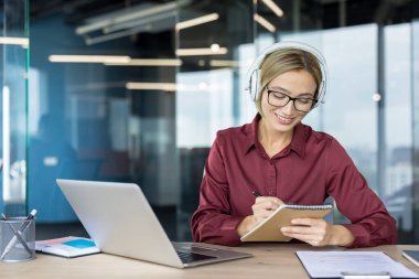 Kulaklık ve gözlük takan bir kadın, masasındaki not defterine notlar yazarken gülümseyen, modern ofis ortamında dizüstü bilgisayarda çalışan, uzaktan öğrenmeye ya da sanal bir konferansa konsantre olan bir kadın.