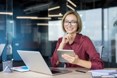 Genç iş kadını güvenle gülümsüyor, elinde dijital tabletle modern bir ofis masasında dizüstü bilgisayarla oturuyor, görevleri yönetiyor ve profesyonelliğini gösteriyor.