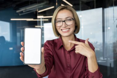 Modern bir ofis ortamında uygulama veya platform sunarken boş beyaz ekranlı akıllı telefonu gösteren genç ve mutlu bir iş kadını.