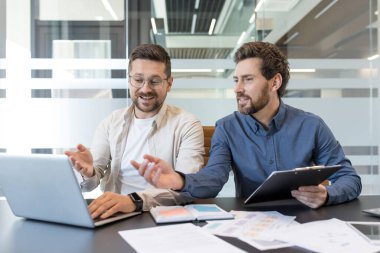 İki işadamı proje ayrıntılarını ve fikirlerini tartışırken bir dizüstü bilgisayar üzerinde birlikte çalışarak modern ofis ortamında takım çalışmalarını ve profesyonel işbirliğini teşvik ediyorlar.