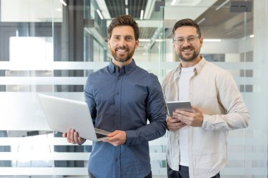 Modern bir ofiste elinde bir dizüstü bilgisayar ve dijital tabletle duran gülümseyen iki iş arkadaşı profesyonel bir ortamda takım çalışmasını ve başarıyı yansıtıyorlar.