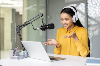 Melez bir kadın podcast kaydediyor, kulaklık takıyor ve dizüstü bilgisayara bakarken profesyonel bir mikrofona konuşuyor, modern çalışma alanındaki içeriği paylaşıyor.