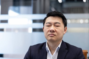 Asyalı işadamı, gözleri kapalı, modern bir ofiste sakin bir meditasyon ve konsantrasyon için bir an duraklıyor, zihinsel refah için farkındalık ve stres rahatlaması uyguluyor.