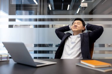 Asyalı işadamı, elleri başlarının arkasında, elleri kapalı bir şekilde ofis koltuğuna yaslanmış, işini bitirdikten ve sakin bir başarı anının keyfini çıkardıktan sonra masadaki dizüstü bilgisayara mutlu bir şekilde gülümsüyor.