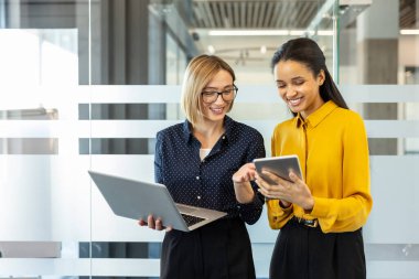 İki gülümseyen iş kadını bir ofiste aktif olarak iş birliği yapıyor ve bir dizüstü bilgisayar ve dijital tablet kullanarak, takım çalışmasını ve modern iletişimi yansıtıyor.