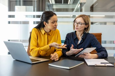 Kurumsal bir toplantı sırasında aktif olarak işbirliği yapan ve iletişim kuran iki farklı iş kadını, biri dijital tablet, diğeri ise el kol hareketi, takım çalışması ve profesyonel tartışmayı vurguluyor.
