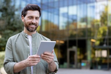 Gülümseyen adam modern bir dijital tabletle etkileşime giriyor, arka planda kentsel cam bir bina ile iletişim ve uzaktan çalışma için kablosuz bir cihaz