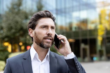 Profesyonel bir iş adamı cep telefonuyla konuşuyor, önemli bilgileri aktarıyor ve çağdaş cam ofis binasının önünde dikiliyor.