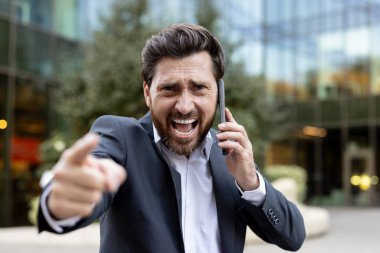 Kızgın iş adamı cep telefonuyla bağırırken bir yandan ileri doğru işaret ediyor. Açık hava ortamında hayal kırıklığını ve saldırganlığı ifade ediyor.