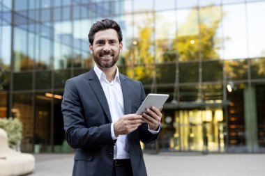 Profesyonel bir işadamı kameraya gülümseyen bir takım elbise ve beyaz gömlek giyerken iki eliyle dijital tablet tutuyor ve modern cam bir binanın önünde dikiliyor.