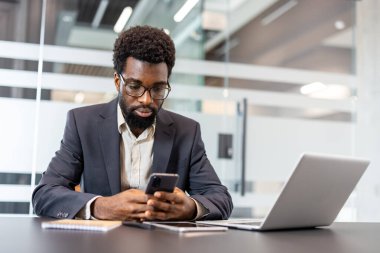 Modern bir masada cep telefonu teknolojisiyle uğraşan genç işadamı, dijital cihazlar arasında çoklu görev yaparken, parlak bir şirket ortamında çalışarak, bağlantı ve üretkenliği sembolize ediyor.