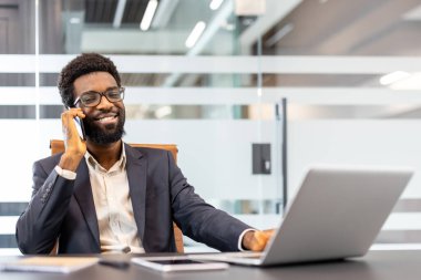 Modern bir ofiste dizüstü bilgisayarla çalışırken cep telefonuyla konuşan gülümseyen profesyonel bir adam net iletişim ve başarılı iş anlaşmaları yapıyor.
