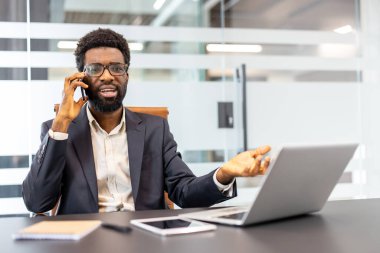 Modern ofisteki masasında önemli bir telefon görüşmesi yapan profesyonel bir adam, iş konseptlerini aktarırken, bilgisayarını ve tabletini kullanarak el kol hareketi yapıyor.