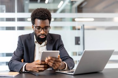 Masasındaki dijital tablete odaklanmış ve kendini adamış genç profesyonel bir adam, modern teknolojiyi çağdaş bir ofis ortamında iş yapmak için kullanıyor.