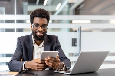 Neşeli erkek profesyonel kameraya bakıyor ve dijital tableti elinde tutarken açık planlı, çağdaş bir çalışma alanında, başarıyı, yönetimi ve küresel iş teknolojisini temsil ediyor.