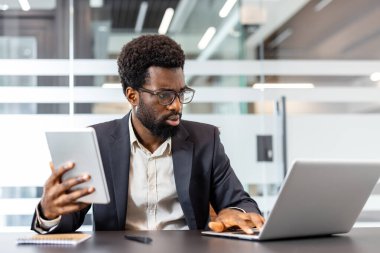 Gözlüklü ve takım elbiseli genç işadamı, elinde dijital tablet, strateji planlaması ve veri analizi yaparken bir dizüstü bilgisayarda yazı yazmaya ve modern bir ofis masasında birden fazla iş yapmaya odaklandı.