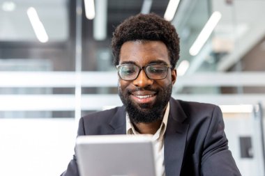 Gözlüklü ve takım elbiseli genç adam, parlak çağdaş çalışma alanında iş iletişimi için dijital tablet kullanırken özgüvenle çalışıyor ve gülümsüyor.
