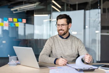 Gözlüklü gülümseyen bir adam masada oturuyor, elinde kalem tutarken bilgisayarına bakıyor ve kağıtları karıştırıyor, modern bir ofiste çalışmalarına odaklanıyor.