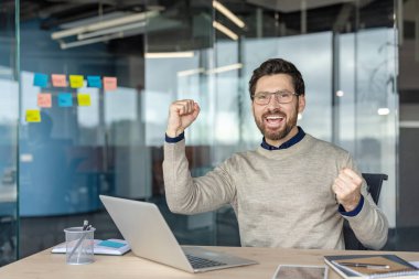 Gözlüklü mutlu sakallı adam modern cam bir ofiste dizüstü bilgisayara yumruk atıyor. Büyük bir iş zaferini heyecanlı, gururlu ve profesyonel tatminle kutluyor.