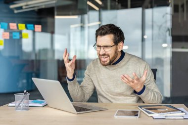 İş adamı stresli ve kızgın hissediyor, bir dizüstü bilgisayara bakarken bağırıyor bir hata mesajı veya problem gösteriyor, modern bir ofiste zor bir sorun yaşıyor