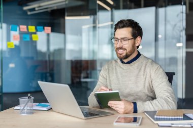 Modern ofisten çalışan mutlu erkek profesyonel, bir proje üzerinde çalışıyor, online bir toplantı sırasında dizüstü bilgisayar ekranına bakarken bir deftere bilgi yazıyor.