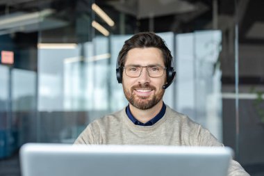 Gözlüklü, kulaklık takan, gülümseyen, modern bir ofiste dizüstü bilgisayarda çalışan, müşteri hizmetleri, online iletişim ve teknik destek kavramlarını tasvir eden bir adam.