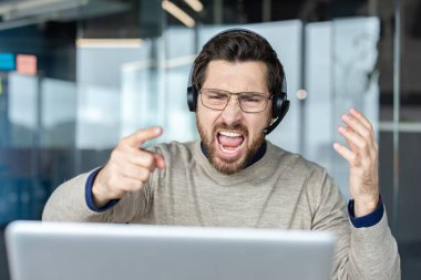 Kulaklık ve gözlük takan, aşırı öfke ve hayal kırıklığını ifade eden, modern ofis ortamında dizüstü bilgisayarda çalışırken ağzını açık gösteren bir adam.