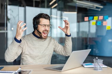 Erkek müşteri hizmetleri görevlisi kulaklık takmış bağırıyor ve el kol hareketi yapıyor. Modern bir ofiste dizüstü bilgisayarda çalışırken kızgınlığını ve hayal kırıklığını ifade ediyor. İşyeri basıncını ve yanmayı simgeliyor