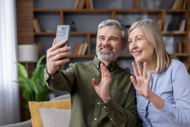 Mutlu son sınıf çifti kanepede oturuyor, video görüşmesi sırasında gülümsüyor ve akıllı telefona el sallıyor, evdeki teknoloji vasıtasıyla aile veya arkadaşlarıyla iletişim kuruyor.