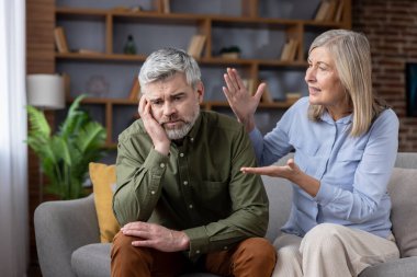 Kanepede oturan yaşlı çift, yüksek sesle konuşan ve el kol hareketi yapan kadın, üzgün ve hayal kırıklığına uğramış görünen erkek, evlerinde ilişki sorunları ve evlilik çatışmalarıyla karşı karşıya.