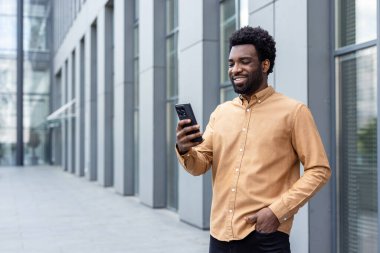 Genç adam dışarıda duruyor, cep telefonunu kullanırken gülümsüyor, çağdaş bir şehir ortamında modern iletişim, iş, teknoloji ve bağlantı kavramlarını yansıtıyor.