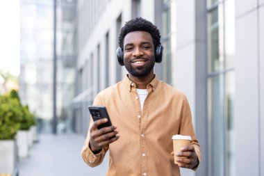 Genç adam gülümsüyor, kablosuz kulaklıkla müzik dinliyor, modern bir şehir ortamında dışarıda yürürken elinde tek kullanımlık bir kahve fincanı ve akıllı telefon tutuyor.