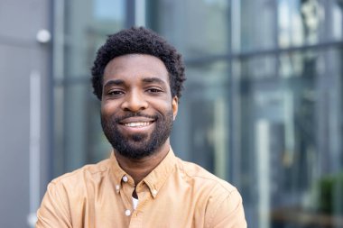 Afro saç stili olan genç işadamı şehir ortamında özgüvenle gülümsüyor, akıllı, sıradan ve ulaşılabilir, başarıyı, liderliği ve parlak bir geleceği tasvir eden bir portre.