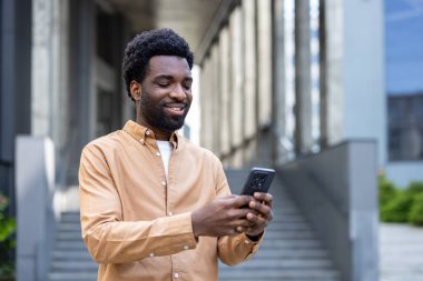 Genç Afro-Amerikalı bir adam bir şehir caddesinde duruyor, akıllı telefonuyla gülümsüyor ve iletişim, sosyal ağ ve modern dijital yaşam tarzını temsil ediyor.