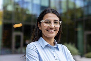 Gülümseyen, berrak çerçeveli gözlük takan ve açık mavi gömlekli genç bir kadın modern cam bir binanın önünde güvenle duruyor, başarıyı ve profesyonelliği temsil ediyor.