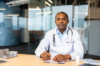 Laboratuvar önlüğü ve steteskop giymiş profesyonel bir erkek doktor modern bir ofiste bir masada oturuyor, bir panoya yazıyor ve kameraya özgüvenle bakıyor, güven ve tıbbi uzmanlığı simgeliyor.