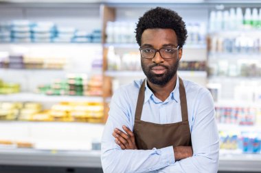 Profesyonel perakende satış elemanı gözlüklü ve önlüklü, bulanık süpermarket raflarının önünde kolları çapraz duran, müşteri hizmetlerini ve işyeri davranışlarını temsil eden