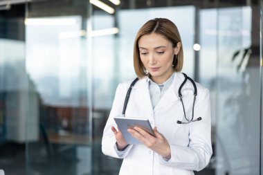 Beyaz önlüklü ve steteskoplu kadın doktor dijital tabletteki tıbbi verileri dikkatle inceliyor, çağdaş bir ofis ortamında duruyor, modern sağlık teknolojisini temsil ediyor.