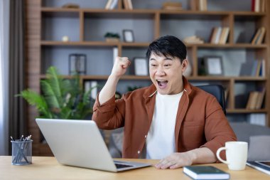 Asyalı bir adam kutlama için elini kaldırıyor ve dizüstü bilgisayarına bakıyor. Başarı, zafer ve mutluluğu ev ofisinden uzaktan çalışarak yaşıyor.
