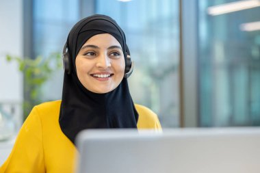 Başörtülü Müslüman bir kadın, tebessüm takıyor, gülümsüyor ve yüzünü çeviriyor. Bu sırada da, modern ve parlak bir ofis ortamında müşterilerine destek ve sanal yardım sunuyor.
