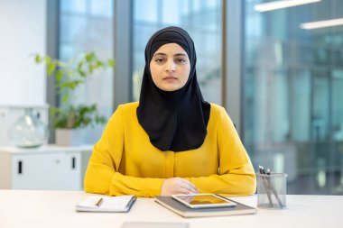 Ciddi Müslüman iş kadını tesettürlü ve sarı gömlekli. Çağdaş ofis ortamında bir masada oturuyor. Profesyonelliği, güçlendirmeyi ve çeşitliliği temsil ediyor.