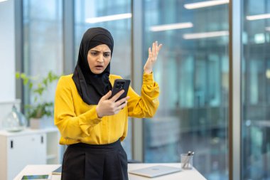 Modern bir ofiste tesettürlü Müslüman bir iş kadını masasında duruyor ve akıllı telefonundan gelen beklenmedik haberlere şok ve hayal kırıklığıyla tepki veriyor.