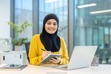 Modern bir ofiste bilgisayarındaki bir toplantıda tesettürlü, tesettürlü, gülümseyen ve not alan Müslüman bir iş kadını.