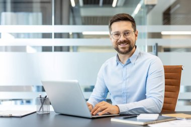 Masasında oturan genç işadamı, modern ofis ortamında dizüstü bilgisayarıyla mutlu bir şekilde çalışıyor, profesyonel başarıyı, teknolojiyi ve kurumsal çevreyi temsil ediyor.