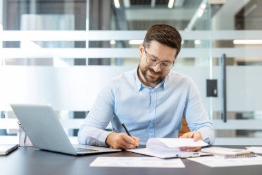 Gülümseyen profesyonel bir adam resmi bir belgeyi kalemle imzalıyor, elinde dizüstü bilgisayarla bir masada oturuyor ve çeşitli evraklarla, verimli mali işleri ve işletme yönetimini temsil ediyor.