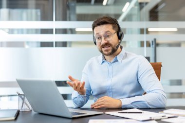 Kulaklık ve gözlük takan işadamı, modern ofis ortamındaki sanal bir toplantı sırasında aktif bir şekilde el kol hareketi yaparak bilgisayarında video konferansı düzenliyor.
