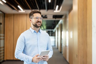Gözlüklü ve mavi tişörtlü gülümseyen işadamı, elinde dijital tablet, profesyonellik ve teknolojiyi temsil ederken çağdaş bir ofis koridorunda güvenle yürüyor.